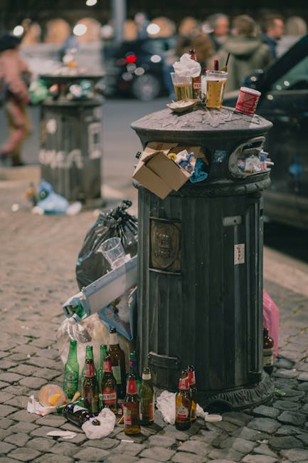A patch of ground beside a grassy slope scattered with various types of waste and discarded packaging materials, including torn and crumpled cardboard boxes, some displaying green and white branding, 