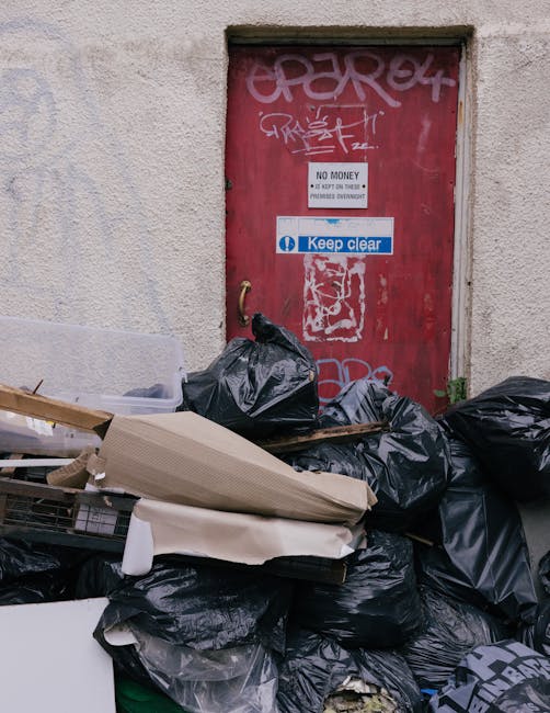 Three black plastic rubbish bags, some with visible white printing, are placed on the edge of a paved sidewalk in front of a dark metal fence with vertical bars. The bags appear to be filled with wast