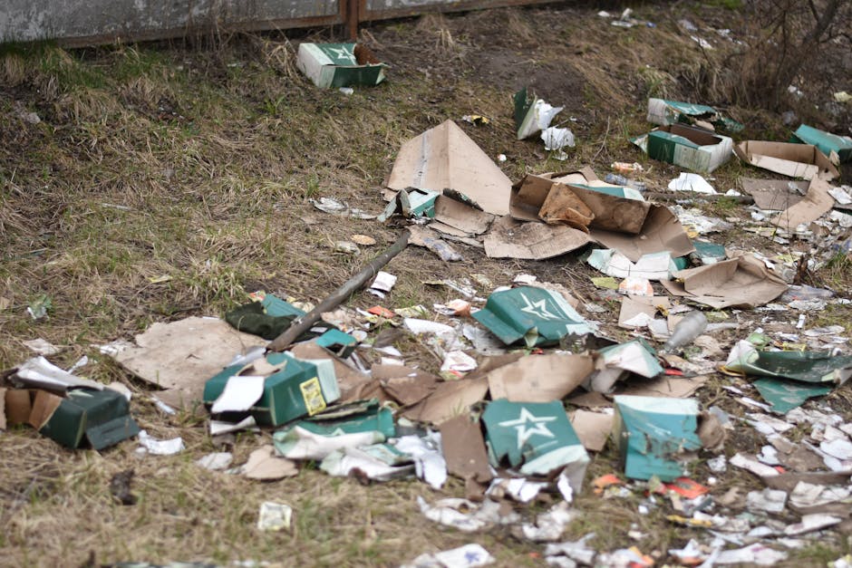 A pile of mixed household rubbish and packaging materials, including flattened cardboard boxes, plastic bags, and paper wrappers, located at the base of an exterior brick wall with a rough concrete fi